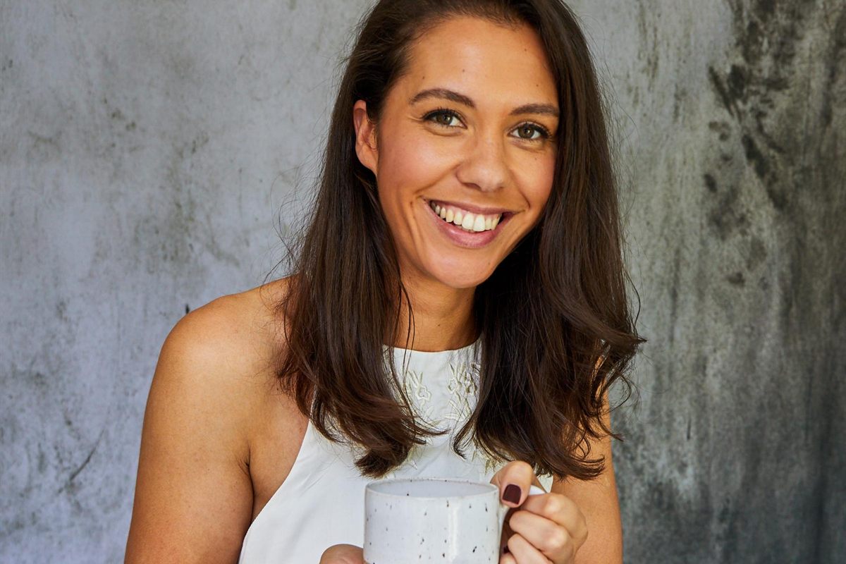 Jade Boswell smiling and holding a speckled ceramic mug of tea against a grey wall
